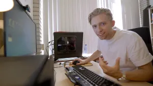 Een foto van presentator Jard. Hij heeft een wit T-shirt aan en zit achter een computer, en vertelt wat in de camera.  