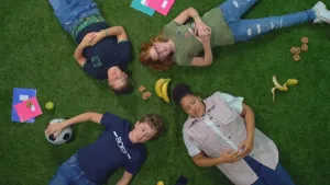Een foto van vier jongeren die op het gras liggen, met de hoofden naar elkaar toe. Je ziet ze recht van boven gefilmd. Op gras liggen ook een voetbal, een banaan, een appel, koeken en wat schriften.