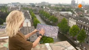 Een foto van presentator Janouk. Ze staan in Amsterdam op het dak van een hoog gebouw. Ze heeft in haar handen een luchtfoto van de Amsterdamse grachtengordel. Onder haar zie je een van die grachten, met oude huizen ernaast.