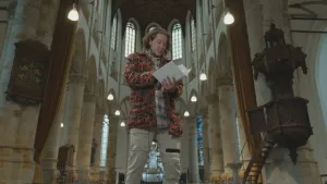 Een foto van presentator Pascal. Hij staat in een grote Middeleeuwse kerk en bladert door een wit boek. 
