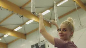 Een foto van presentator Janouk in een sporthal. Ze houdt een trapeze vast en kijkt in de camera. 