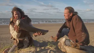 Een foto van presentator Pascal. Hij zit samen met een andere man op een strand en heeft een groot bot in zijn handen. Het is een bot van een mammoet. Hij kijkt er vol bewondering naar.