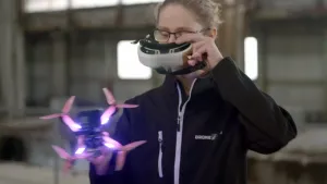 Een foto van een vrouw in een grote hal. Ze heeft een drone vast in een hand en de bijbehorende FPV-bril in haar andere hand. Ze zet die bril op of af. 