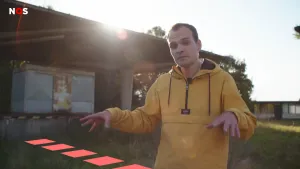 Een foto van een presentator met een gele jas op een grasveld. De presentator houdt zijn handen van elkaar af met de handpalmen naar beneden en gestrekte vingers. Vóór de foto en de presentator staan vijf getekende, rode vierkanten in één lijn, een grens.