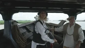 Een foto van presentator Janouk in een heel oud houten schip. Ze heeft Middeleeuwse kleren aan en staat aan het roer. Naast haar staat een man, ook in Middeleeuwse kleren. Ze varen op een rivier.