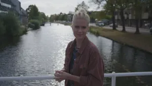 Een foto van presentator Janouk. Ze staat in een stad op een brug en kijkt in de camera. Achter haar zie je water en huizen. 