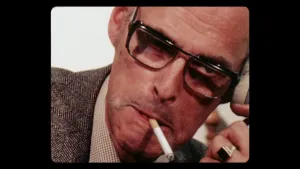 Een foto van een man die een sigaret in zijn mond heeft. Hij draagt een zwarte bril, een donkergrijs pak en een zegelring, goudkleurig met een zwarte rechthoek erop. Rechts is nog net de hoorn van een ouderwetse, vaste telefoon te zien aan zijn linkeroor.