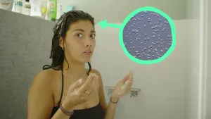 Een foto van Manon in een zwart badpak in een douche. Ze kijkt naar de camera met haar handpalmen omhoog voor haar lichaam. Ervoor staat een lichtgroene cirkel met pijl die naar haar hoofd wijst met een grijze achtergrond met kleine, witte rondjes erin.