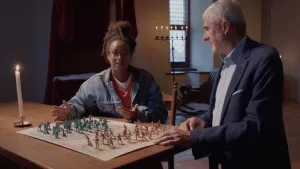 Een foto van presentator Eva. Ze is in Slot Loevestein en zit aan een houten tafel. Op die tafel ligt een oude landkaart. Daarop staan groepen mannetjes in twee verschillende kleuren. Dat zijn twee legers. Naast Eva staat een man. Hij kijkt naar haar.