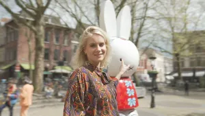 Een foto van presentator Janouk die buiten op een plein staat en wijst naar een beeld van Nijntje.