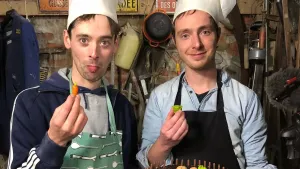 Een foto van Pieter en Willem in hun werkplaats. Ze hebben koksmutsen op en kijken allebei in de camera. Willem houdt een marsepeinen worteltje in zijn handen en Pieter een stukje groene groente, ook van marsepein.