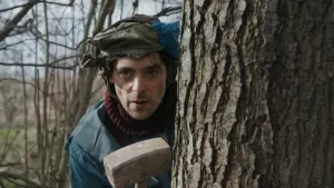 Een foto van Willem in een bos. Hij kijkt langs een boom en loert ergens op. Hij heeft een houten hamer in zijn handen.