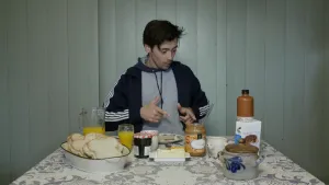 Een foto van presentator Willem die aan een tafel zit waar brood en broodbeleg op staat. Waaronder een pot pindakaas en een pot jam. Ook staat er een kan met sinaasappelsap en een glas op tafel. Willem wijst naar iets wat op tafel ligt.