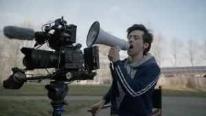 Een foto van presentator Willem die in een stoel zit en in een megafoon praat. Er staat een grote video camera naast hem. Een megafoon is een soort luidspreker waar je hard door kunt praten.