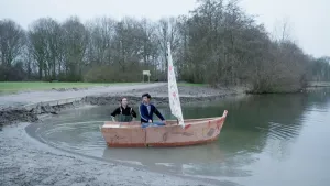 Een foto van presentator Willem die aan de oever in een klein zeilbootje zit. Presentator Pieter staat in het water naast het zeilbootje.