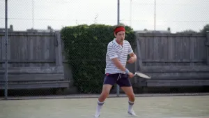Een foto van presentator Willem op een tennisbaan. Hij heeft tenniskleren aan en een rode hoofdband om zijn hoofd. Hij heeft net een bal geslagen. 