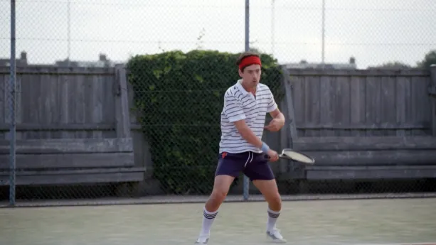 Een foto van presentator Willem op een tennisbaan. Hij heeft tenniskleren aan en een rode hoofdband om zijn hoofd. Hij heeft net een bal geslagen. 