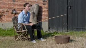 Een foto van presentator Pieter. Hij zit op een houten stoel voor een schuur. Hij vist met een zelfgemaakte hengel in een ronde ijzeren bak. 