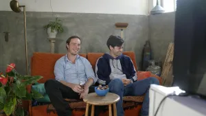 Een foto van de presentatoren Pieter en Willem. Ze zitten naast elkaar op een oranje bank en kijken TV. Pieter heeft een blikje bier in zijn handen. Ze lachen allebei. Voor hen staat een krukje met daarop wat koekjes.