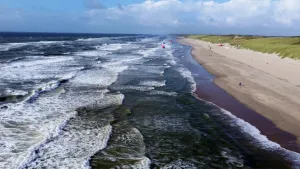 Een foto van een kustgebied, waar links witte golven in het water te naar het zandstrand stromen. Er lopen enkele mensen over het strand en twee mensen in de verte hebben een kleurrijke vlieger  vast. Rechts is een stukje van de groene duinen te zien.
