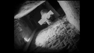 Een zwart-wit foto vanaf de bodem van een rechthoekig gat dat op de niet-zichtbare tunnel onder de Berlijnse muur uitkomt. Er hangt een dun touw naar beneden in het gat en op de houten plank erboven staat een witte zak en steken benen van een mens uit.