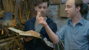 Een foto van de presentatoren Pieter en Willem in hun werkplaats. Willem heeft een boek open. Pieter heeft een bitterbal in zijn handen. Beiden kijken naar de bitterbal. 