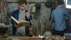 Een foto van de presentatoren Pieter en Willem in hun werkplaats. Willem leest voor uit een groot boek. Pieter staat op de achtergrond. Je ziet hem op de rug.