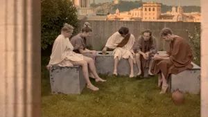 Een foto van vijf mannen en vrouwen die gezamenlijk zitten te poepen. Ze hebben oude Romeinse kleren aan. Op de achtergrond zie je een oude Romeinse stad.