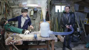 Een foto van de presentatoren Willem en Pieter in hun werkplaats. Pieter heeft een winterjas aan en houdt een grote doorzichtige buis tegen zijn jas. Willem is bezig met een bladblazer die ook is aangesloten op die buis. Er komt veel dons uit de buis.