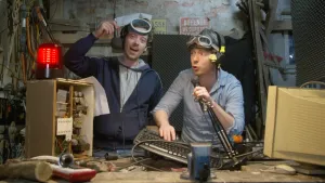 Een foto van de presentatoren Pieter en Willem in hun werkplaats. Beiden hebben een microfoon vast en koptelefoons op. Links in beeld zie je een rode lamp die brandt. Ze maken radio.