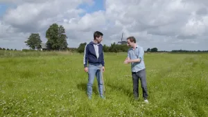 Een foto van de presentatoren Willem en Pieter in een groot weiland. Ze kijken naar elkaar. Willem staat bij een schep die in de grond gestoken is. Op de achtergrond zie je een boerderij en een molen.