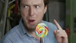 Een foto van presentator Pieter. Hij heeft een grote kleurrijke lolly in een hand en houdt de wijsvinger van zijn andere hand omhoog. Hij is iets aan het vertellen.