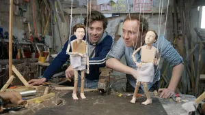 Een foto van twee mannen, Pieter en Willem. Ze staan in een werkplaats en kijken naar twee houten marionet-poppen van zichzelf. Die staan op een tafel, en hangen aan touwtjes. 