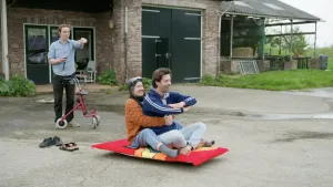 Een foto van presentator Willem die buiten voorop een rijdend vliegend tapijt zit. Achter hem zit er een vrouw op het vliegen tapijt met haar armen om Willem heen. Presentator Pieter staat op de achtergrond met een afstandsbediening in zijn hand.