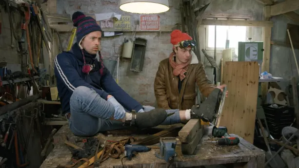 Een foto van presentatoren Willem en Pieter die in een schuur zijn. Willem zit op tafel met een paar schaatsen aan.