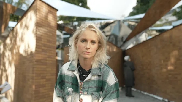 Een foto van presentator Janouk. Ze kijkt met een serieuze blik in de camera. Ze staat voor enkele grote muren die je onscherp op de achtergrond ziet. Een persoon staat van dichtbij naar die muur te kijken. 