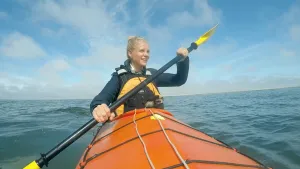 Een foto van presentator Janouk die in een kajak op zee aan het varen is. Een kajak is een kleine een persoons boot waar vooral mee op rivieren wordt gevaren.