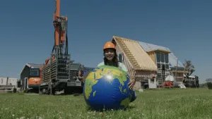 Een foto van een presentator die buiten zit met een opblaasbare wereldbol voor haar. Op de achtergrond wordt er een huis gebouwd en staan er verschillende bouw machines.