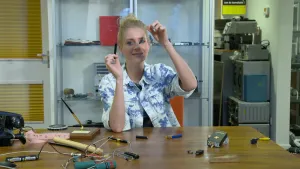 Een foto van presentator Janouk. Ze zit aan een bureau en heeft een deel van een pen vast. Ze toont dat aan de kijker. Op het bureau liggen allerlei spullen zoals een pakje sigaretten, batterijen en een aansteker.