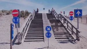 Een foto van een brede houten trap op het strand, de duinen in. Voor die trap staan allerlei verkeersborden om te ervoor te zorgen dat je rechts blijft lopen en anderhalve meter afstand tot andere mensen houdt. 