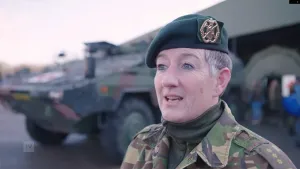 Een foto van een vrouwelijke soldaat. Op de achtergrond is er een leger auto te zien. 