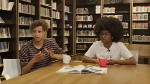 Een foto van twee vrouwen die aan een tafel zitten in een bibliotheek. Op tafel ligt er een open tijdschrift.