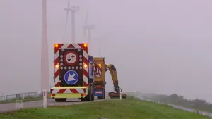 Een foto van een weg met daarop twee vrachtwagens. De voorste vrachtwagen heeft een arm waarmee het gras langs de weg wordt gemaaid. Op de achtergrond zie je auto's rijden en je ziet enkele grote windmolens.