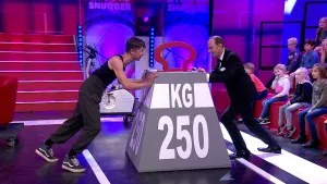 Een foto van twee mannen die allebei aan één kant van een groot gewicht staan en ertegen duwen. Op het grijze gewicht met rood hengsel eraan staat de tekst "250 KG" of kilo. De één draagt sportschoenen en een zwart hemd, de ander draagt een zwart pak.