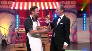 Een foto van twee mannen voor een marktraam met een rood en wit doek erop en vol met dozen lichtbruine eieren. Eén van de mannen heeft een wit schort aan en een rieten mand vast vol gekleurde paaseieren. De andere man houdt een onbeschilderd ei omhoog.