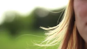 Een foto met rechts een klein stukje van de wang van een persoon voor een onscherpe, groene natuur. Links is het haar van de persoon te zien, dat naast het gezicht naar beneden hangt. Een paar plukken van het blonde haar waaien iets omhoog door de wind. 
