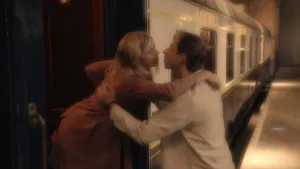 Een foto van een vrouw in de deuropening van een trein en een man op het perron. De vrouw heeft haar arm op de schouder van de man liggen, de man zijn handen op de rug van de vrouw. Ze reiken naar elkaar toe, om elkaar een kus op de mond te geven. 