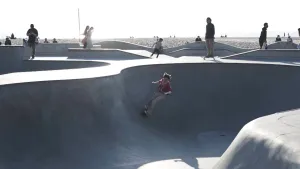Een foto van een grijze skatebaan, waarin iemand in een rood shirt en lichtbruine broek op een skateboard van de ronde, holle zijkant afrolt. Aan de kant staat iemand op een skateboard en iemand met een skateboard loopt weg. Erachter ligt een zandstrand.