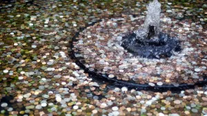 Een foto van een fontein waar in het midden een straal water omhoogspuit. In de binnenste, zwarte ring van de fontein en verder naar de niet-zichtbare zijkant ligt de fontein vol met muntgeld van verschillende kleuren en groottes.