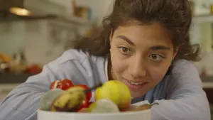 Een foto van een presentator die kijkt naar fruit in een witte schaal: appels, een banaan, tomaten en een sinaasappel. Op twee stuks fruit zijn groenwitte vlekken te zien. Dit is schimmel, dat komt op eten te zitten dat bedorven en niet meer eetbaar is. 